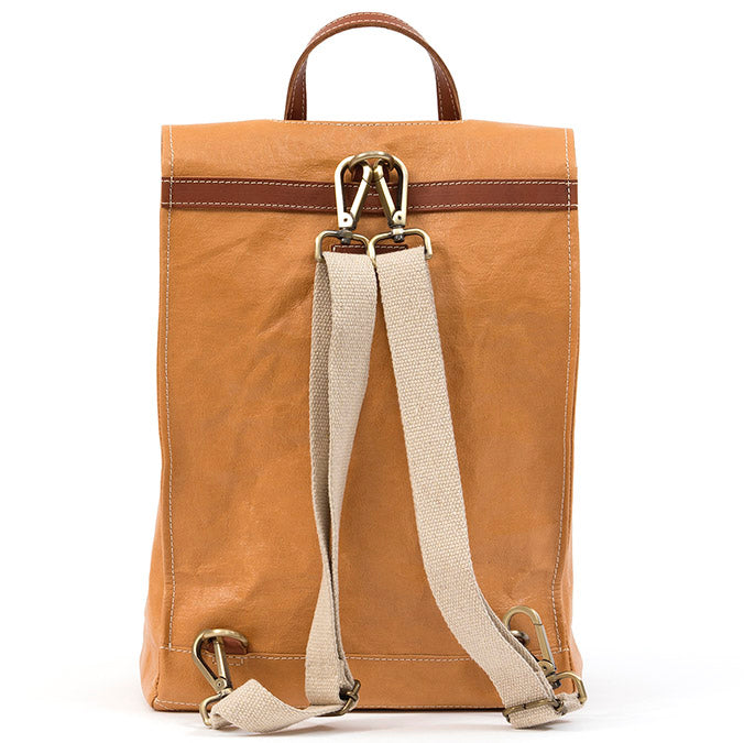 A tan washable paper backpack is shown from the back angle. It features white stitching, a brown strip across the back, a brown top handle and cream cotton canvas straps with gold metallic hardware.