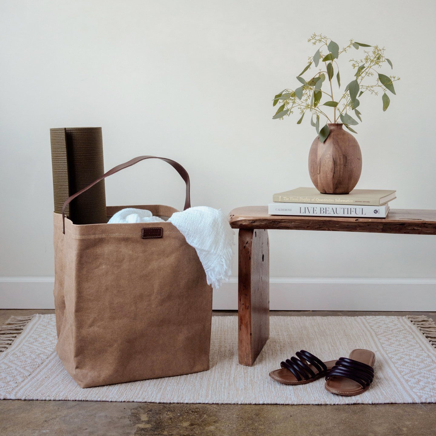 A natural tan washable paper handbag sits in a home setting, containing a towel and a yoga mat. At right sits a wooden bench, a wooden vase containing leaves, and a pair of flat sandals.
