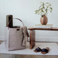 A grey washable paper handbag sits in a home setting, containing a towel and a yoga mat. At right sits a wooden bench, a wooden vase containing leaves, and a pair of flat sandals.