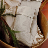 A tan washable paper basket is shown holding two washable paper Christmas stockings, one large and one small. The stockings are pale cream in colour with a platinum metallic cuff, heel and toe.