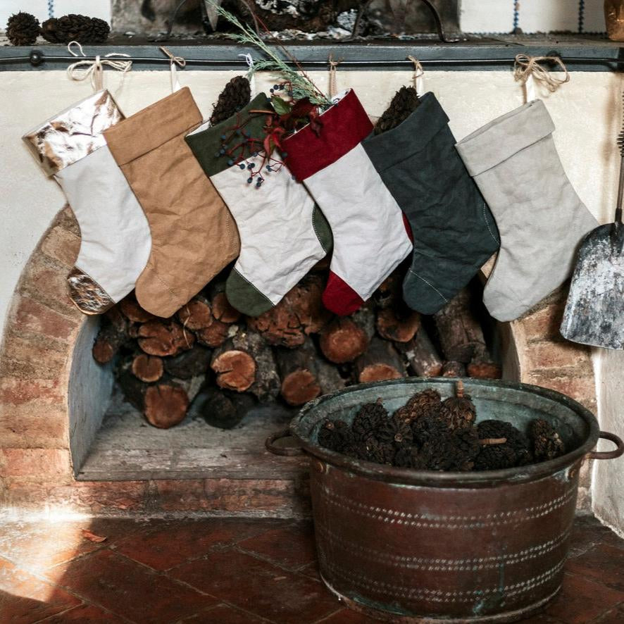 A stone fireplace is shown with six washable paper Christmas stockings hung in a row. In front of the fireplace is a metal bucket containing pine cones.