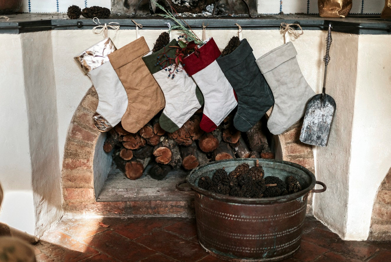 A stone fireplace is shown with six washable paper Christmas stockings hung in a row. In front of the fireplace is a metal bucket containing pine cones.