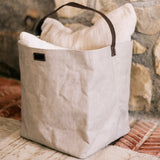 A grey washable paper bag with a singular brown strap sits in a home setting, containing cushions.