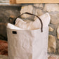 A grey washable paper bag with a singular brown strap sits in a home setting, containing cushions.