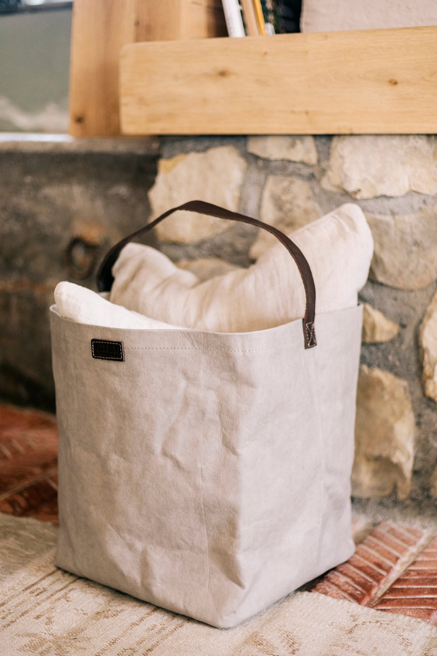 A grey washable paper bag with a singular brown strap sits in a home setting, containing cushions.