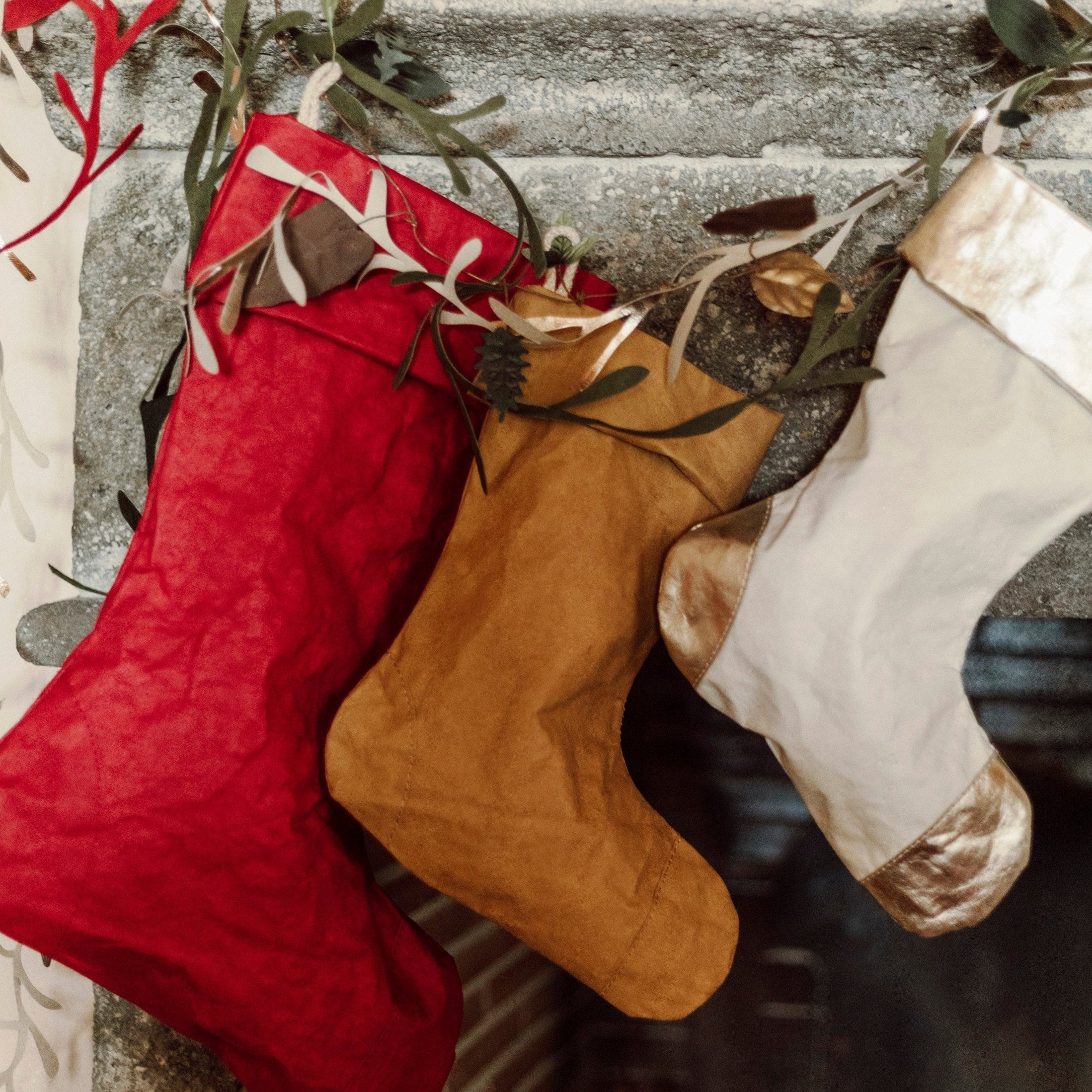 Varying washable paper garland wreaths are shown in beige, green and red atop a mantlepiece containing three washable paper Christmas stockings.