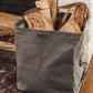 A washable paper bag is shown in grey with a singular strap. It sits at the side of a fireplace, containing logs.