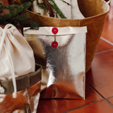 A gold metallic washable paper gift pouch sits in front of a tan washable paper tote bag, under a Christmas tree.