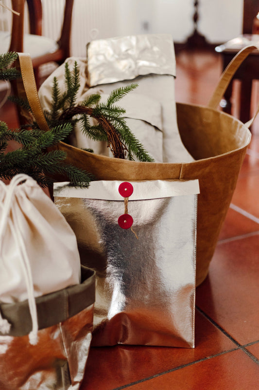 A gold metallic washable paper gift pouch sits in front of a tan washable paper tote bag, under a Christmas tree.