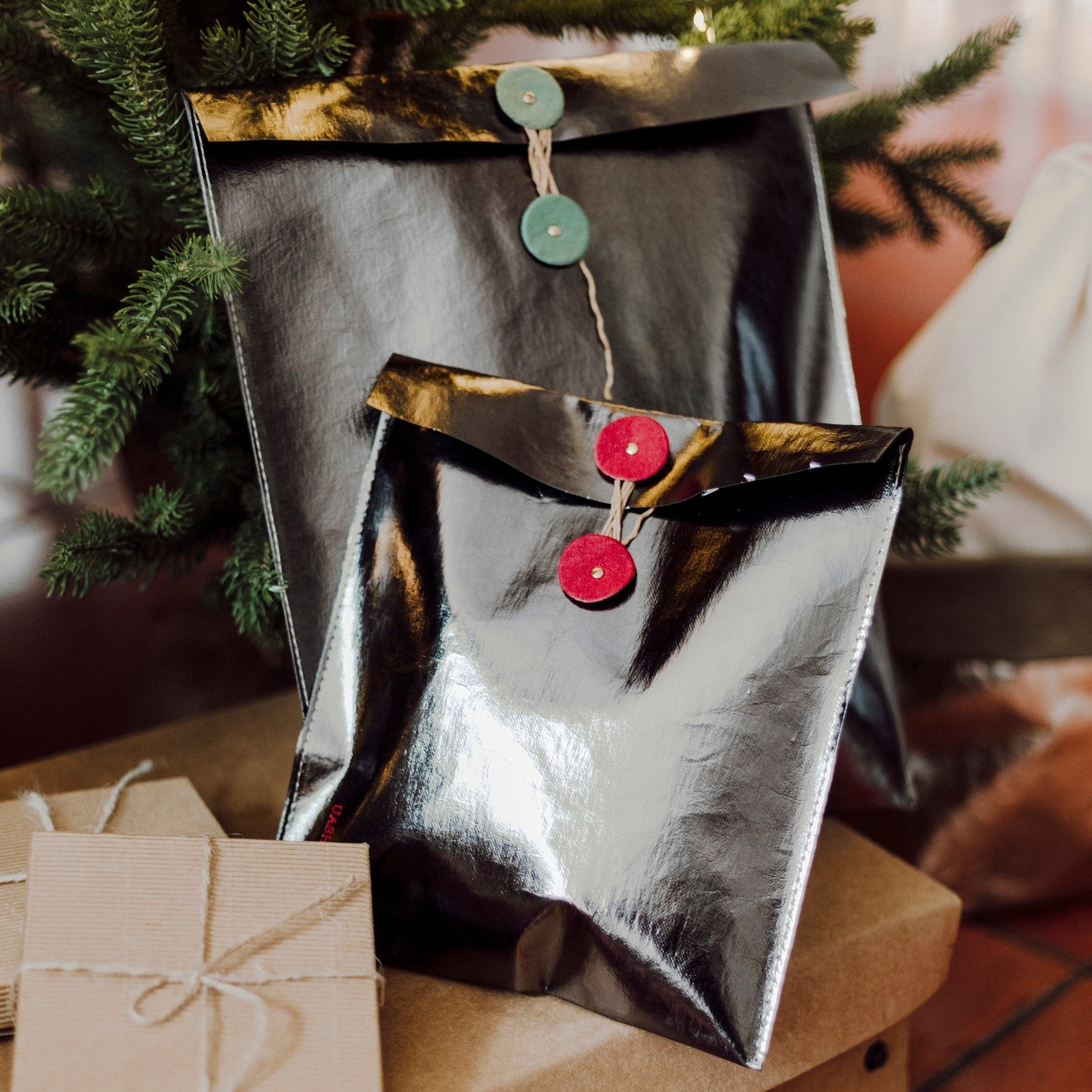 Two pewter metallic gift pouches with twine closures (one red, one green) sit in front of each other atop a washable paper lidded box. A Christmas tree is at the rear of the image.