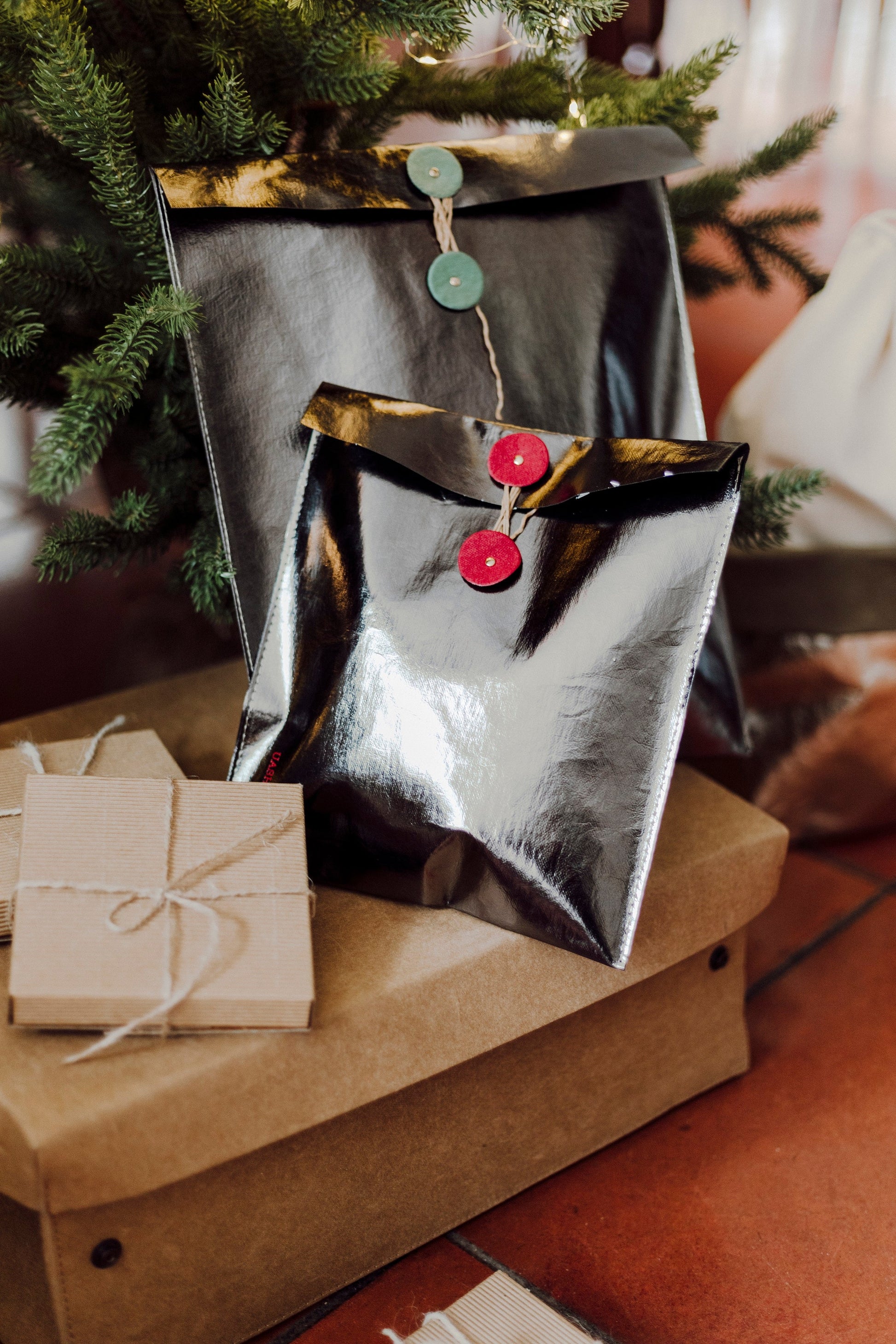 Two pewter metallic gift pouches with twine closures (one red, one green) sit in front of each other atop a washable paper lidded box. A Christmas tree is at the rear of the image.