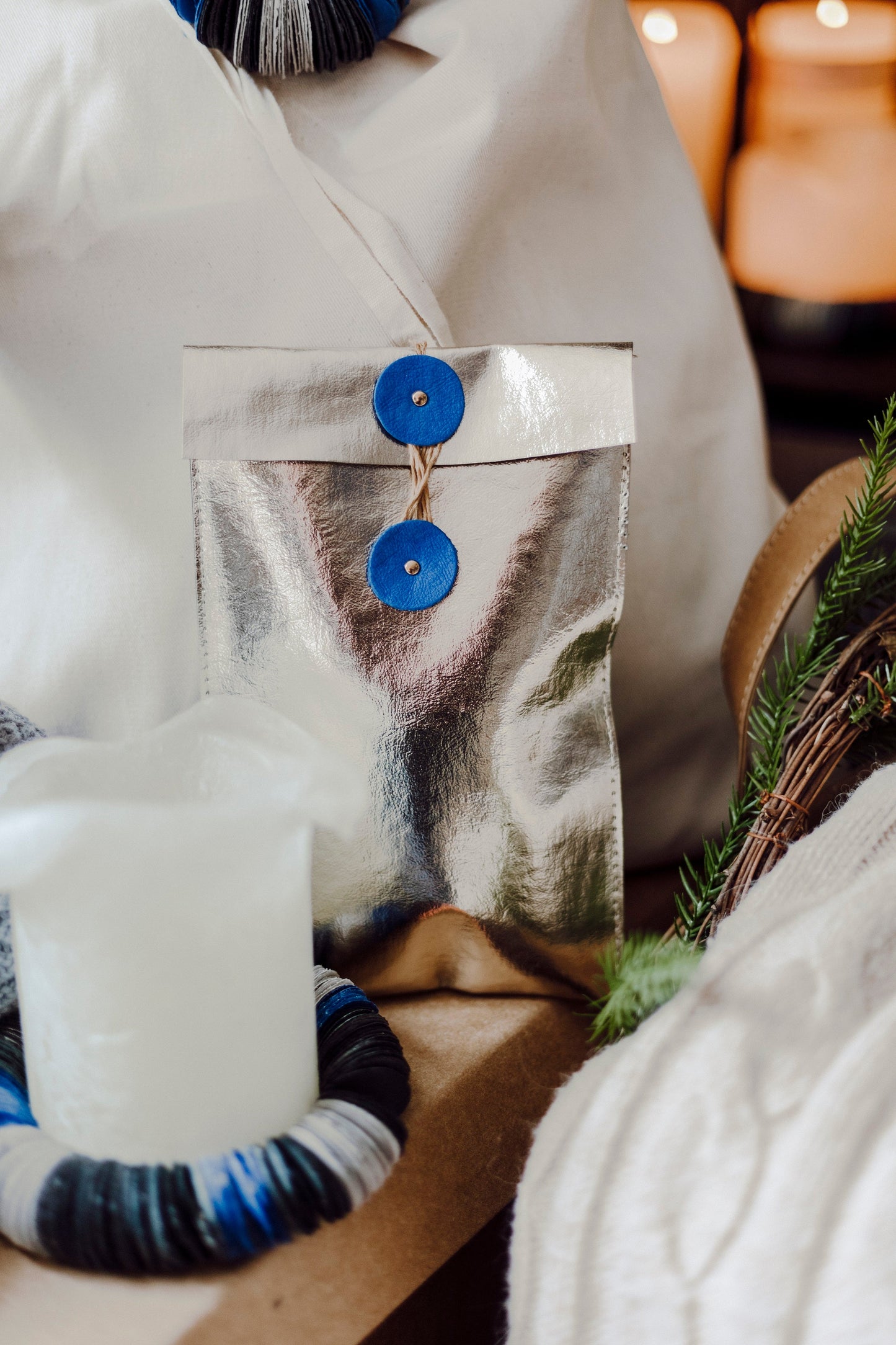 A metallic gold gift pouch featuring a twine closure with blue washable paper discs sits in a home setting. Also visible are two washable paper rings, one around a pillar candle and the other tied to a cotton sack.