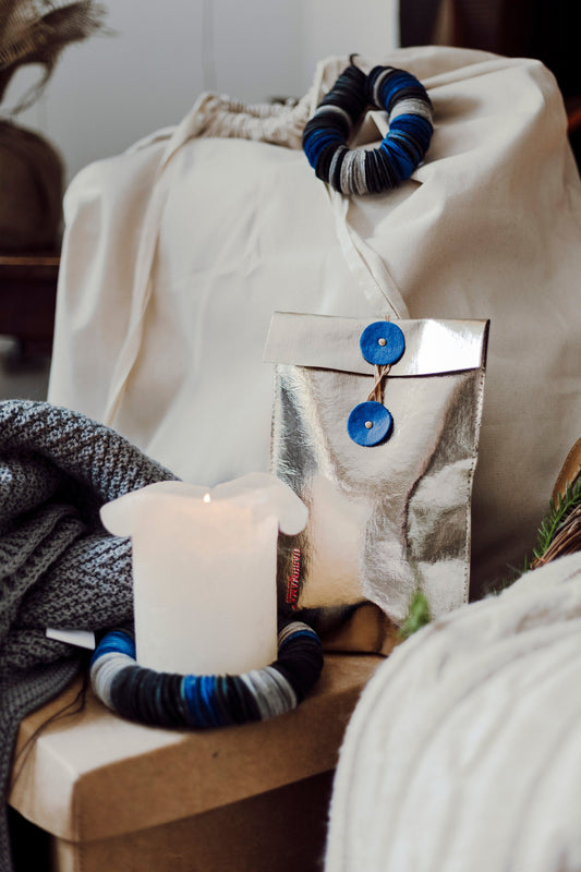 A metallic silver gift pouch featuring a twine closure with blue washable paper discs sits in a home setting. Also visible are two washable paper rings, one around a pillar candle and the other tied to a cotton sack.