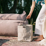 A woman is shown barefoot outdoors next to an outdoor sofa. In her hand rests a grey washable paper handbag with a singular brown strap.