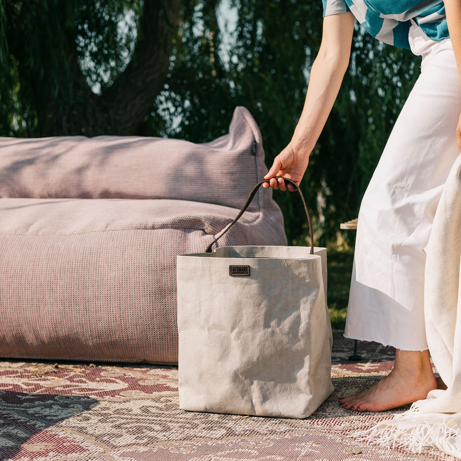 A woman is shown barefoot outdoors next to an outdoor sofa. In her hand rests a grey washable paper handbag with a singular brown strap.