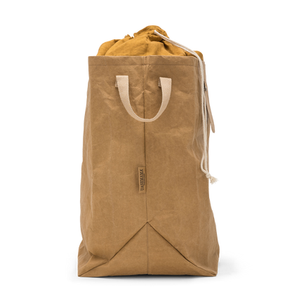 A washable paper laundry bag is shown in tan from a side angle. It features two side handles, a popper tab, and an interior drawstring lining.