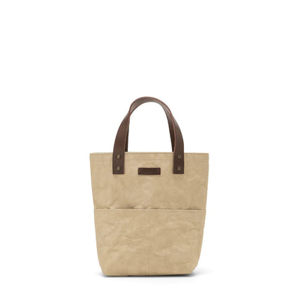 A sand coloured washable paper tote bag with two short brown leather handles and external pockets