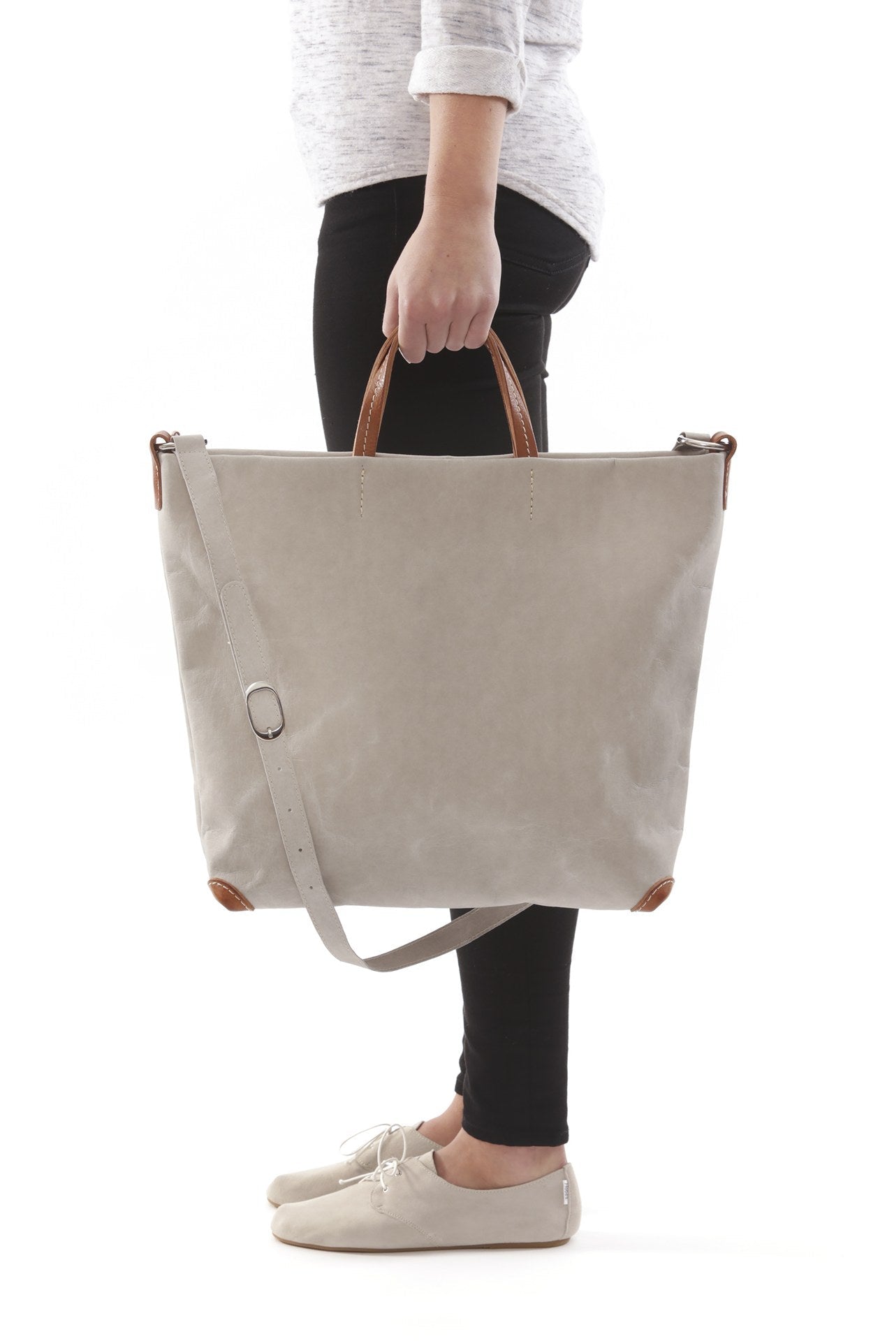 A woman is shown standing, and from a side view. She is carrying a grey washable paper tote bag by short leather handles. The bag's long strap is shown draped over the bag.