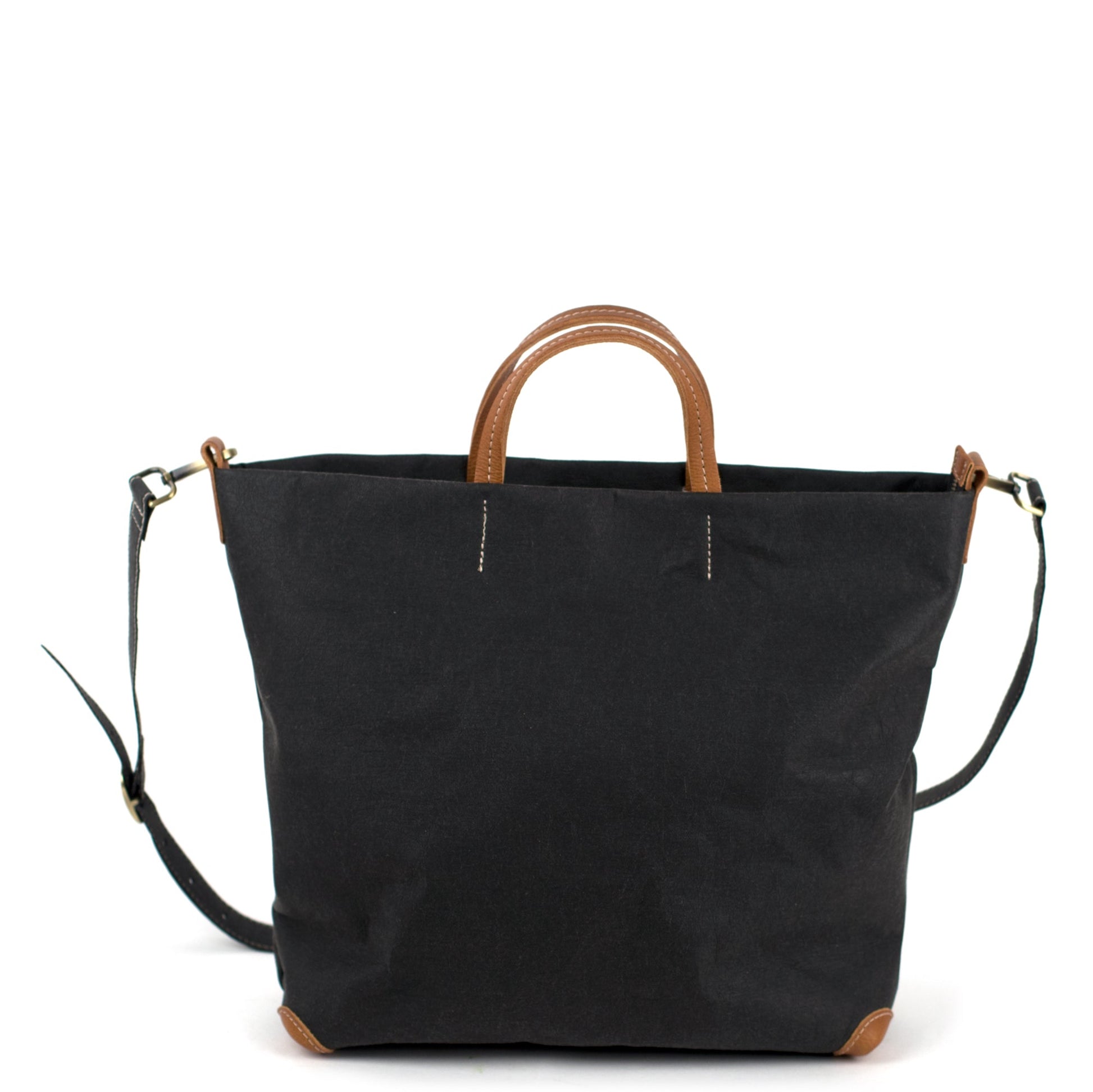 A square black washable paper tote bag is shown with one long black shoulder strap and two small tan handles. The corners of the bag are protected by brown corners.