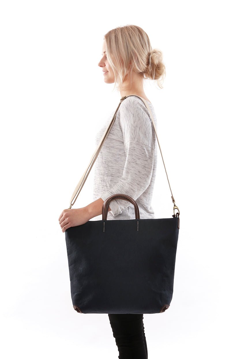 A woman is shown from a side view. She is carrying a black washable paper tote bag on her shoulder by a long strap. Her hand rests on the top of the bag.