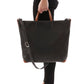 A woman is shown standing, and from a side view. She is carrying a black washable paper tote bag by short leather handles. The bag's long strap is shown draped over the bag.