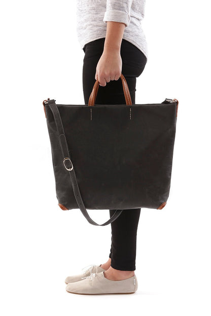 A woman is shown standing, and from a side view. She is carrying a black washable paper tote bag by short leather handles. The bag's long strap is shown draped over the bag.