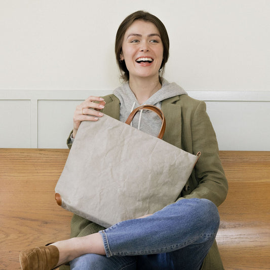 A woman is shown sitting on a wooden bench. She is holding a grey washable paper tote bag.