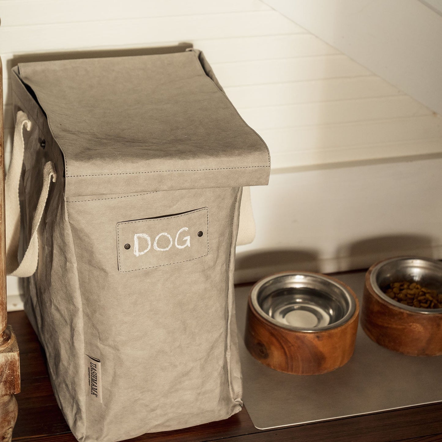 A washable paper recycling bag in pale grey is seen standing next to two wooden and metal dog bowls. One bowl contains water and the other contains dog food. The washable paper tag on the washable paper recycling tote reads "DOG".