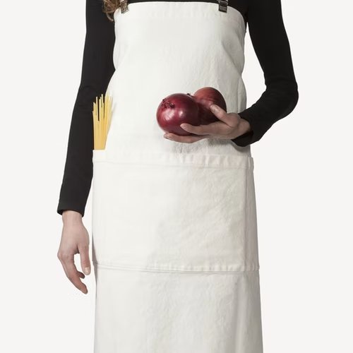 A woman is shown standing holding 2 red onions. She is wearing a beige cotton apron with washable paper braces. Spaghetti is shown in the front pocket of the apron.