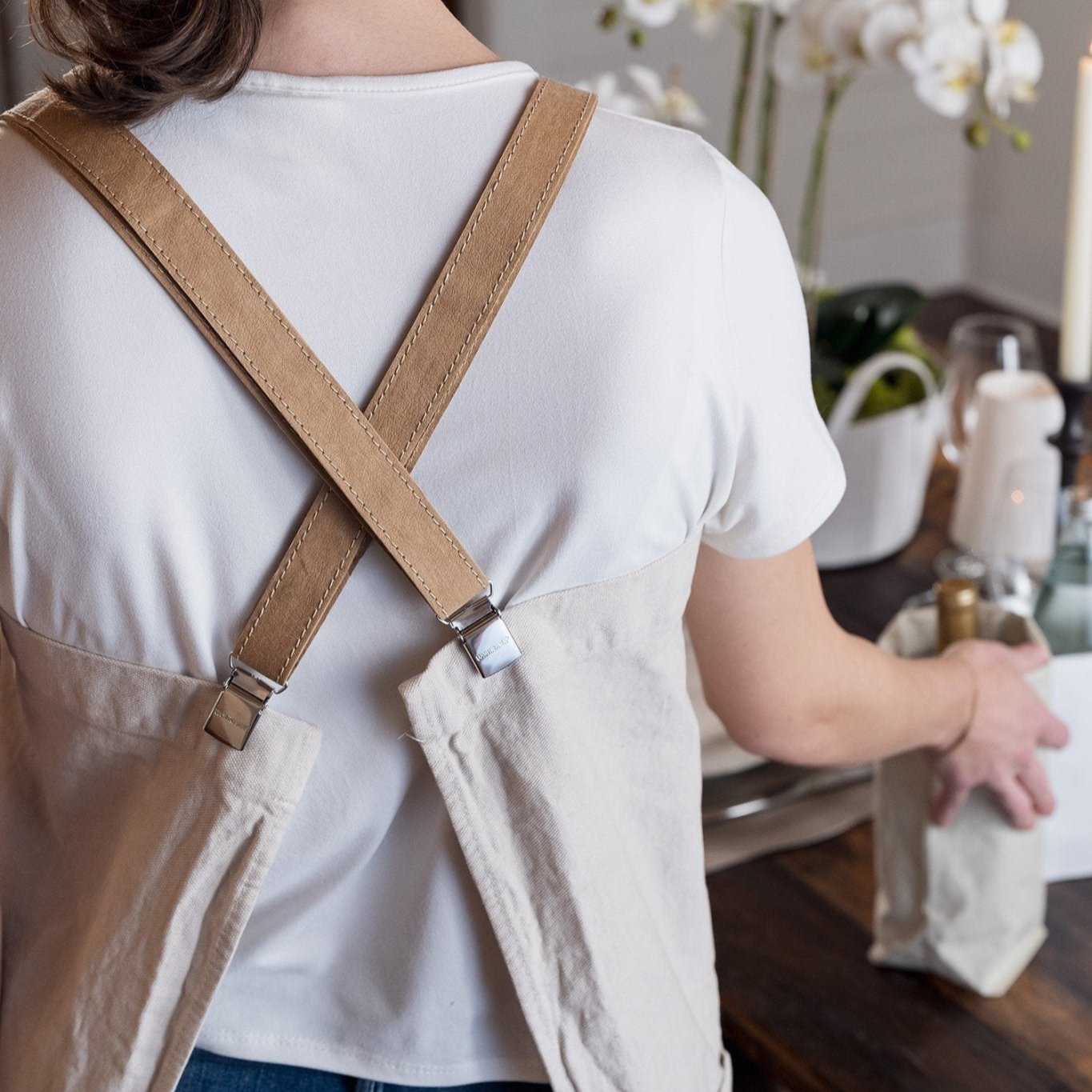 A woman is shown standing with her back to the camera. She is wearing a cotton apron which fastens at the back with washable paper braces. They cross over her back. She is shown putting a washable paper wine bag on a dining table.