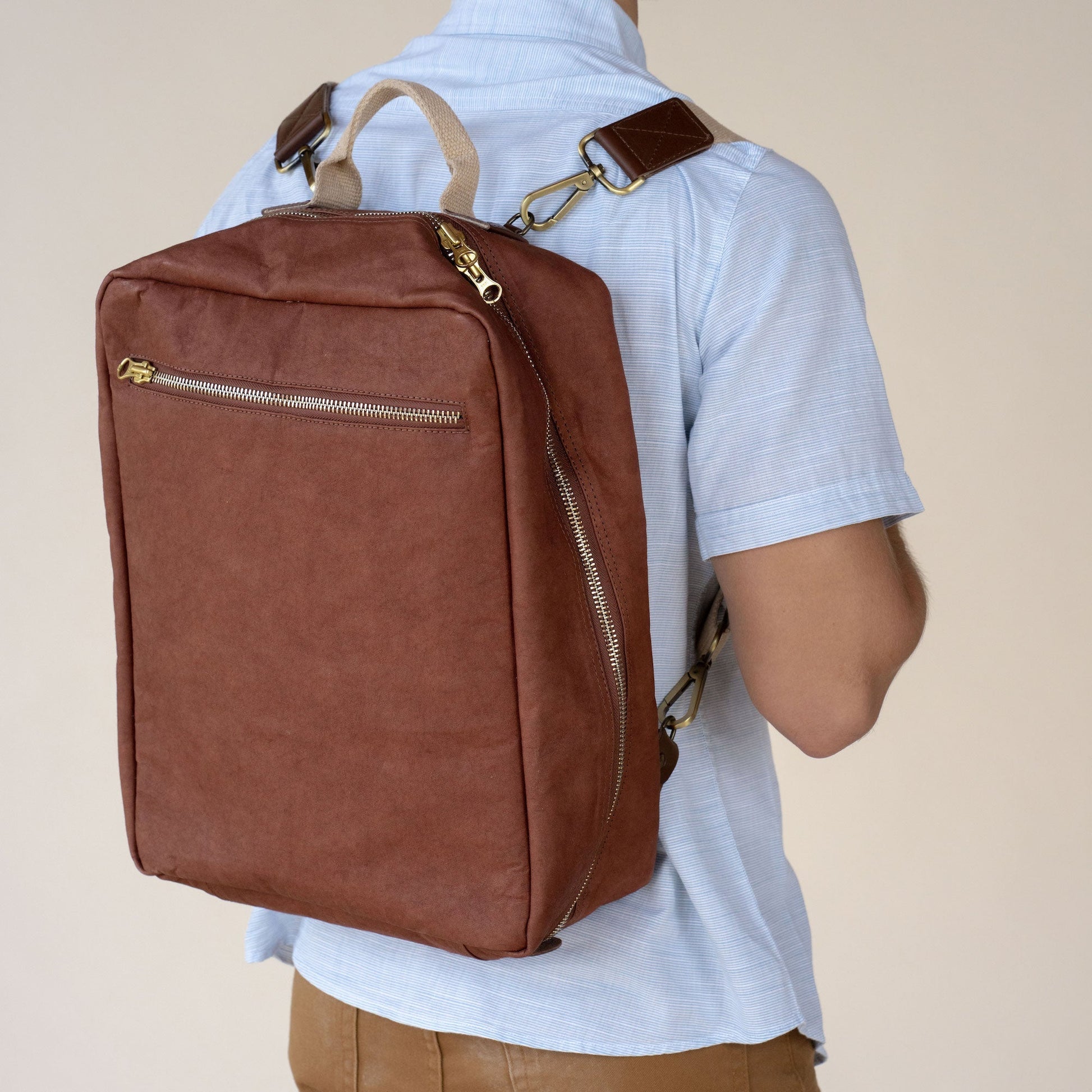 A man is shown standing wearing a square shaped washable paper backpack in a congac colour.
