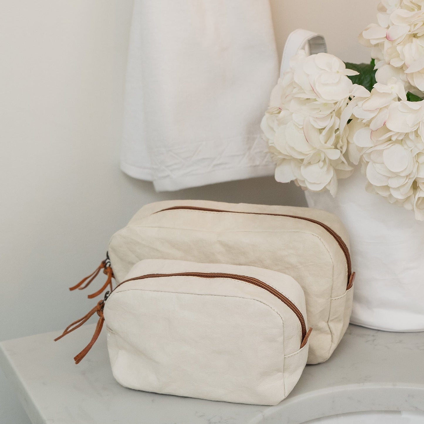The image shows two white washable paper toiletery bags, in medium and large. The bags are closed and are sitting in front of a white washable paper bucket containing white flowers. Hanging on the wall is a white towel.