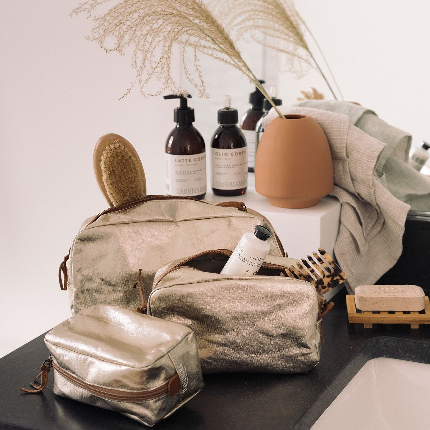 A set of three platinum metallic washable paper toiletry bags is shown. One contains a hairbrush, and another contains a comb and a tube of hand cream. Also shown are bottles of beauty products and a terracotta vase with a sheaf of wheat.