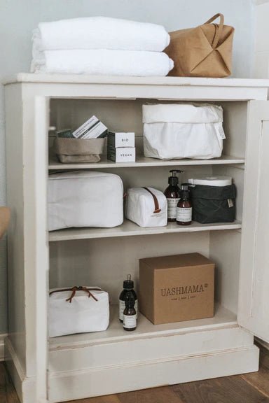The image shows a three shelf bathroom storage unit. On the top are two folded towels and a brown washable paper toiletry bag. On the shelves are an assortment of washable paper toiletry bags in white, washable paper bags in dark grey and grey and some beauty products.