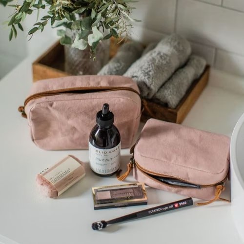 The image is from a bathroom. There are two pale pink washable paper toiletry bags, one of which is lying down. Both are open. Around the bags are a selection of skincare and make up products. In the background is a wooden tray containing rolled up towels and a plant.