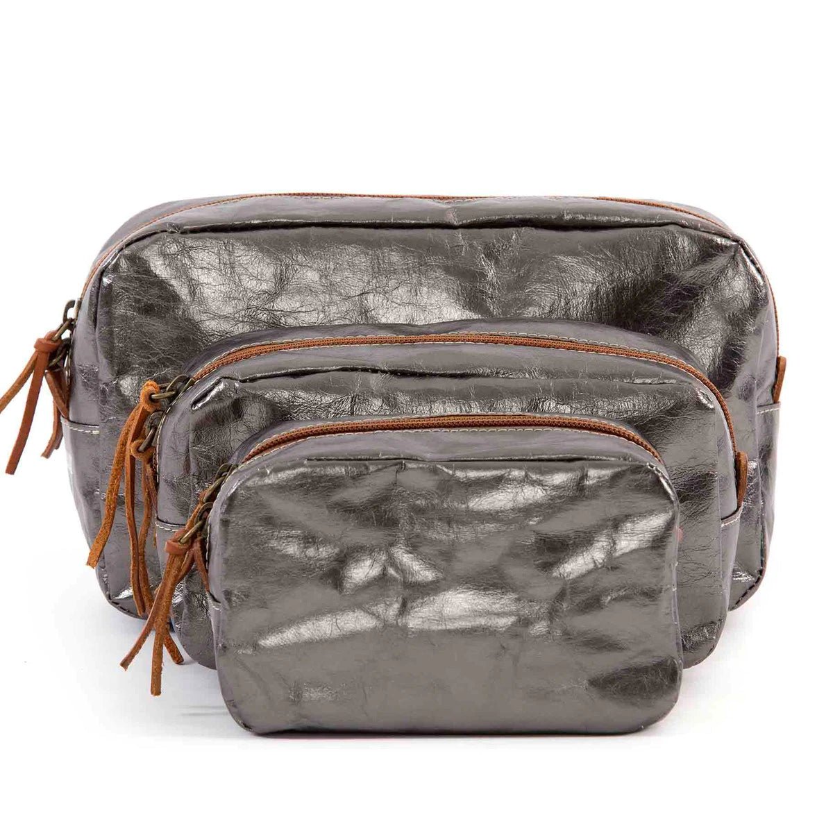 A set of three washable paper toiletry bags is shown. Each bag has a brown zip with a tan leather zip pull. The smallest bag is at the front, then the medium bag and the large size at the back. The bags shown are dark grey metallic in colour.