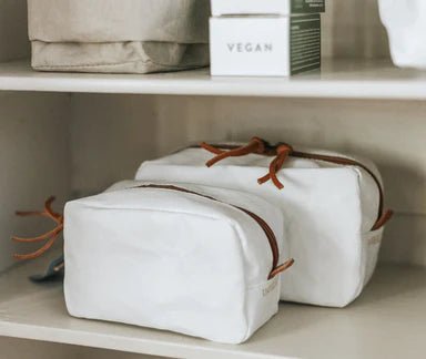 The image shows two shelves. On the top shelf is a washable paper tray in grey containing two boxes of soap. On the bottom shelf are two washable paper toiletry bags in white.