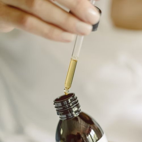 The image shows a dropper being filled from a dark brown bottle of body oil.