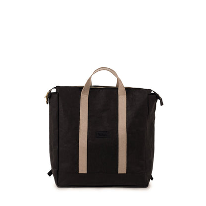 A black washable paper backpack is shown from the front. Square in design, it features a top zip and two cream canvas handles at the front and at the back.