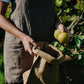 A woman wearing a brown linen and washable paper apron holds a washable paper lunch bag in her hand. Her right hand is putting lemons inside the bag. 