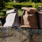 Two washable paper lunch bags sit atop an outdoor wall, next to a terracotta plant pot. The bag at left is grey with a rolled closure, the bag at right is in a natural tan colour with the top rolled down.