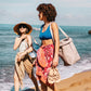 Two women walk along a beach, each carrying a bag. The woman at left wears an orange sun hat, beige swimsuit and sarong, and carries a gold metallic washable paper tote bag. The woman at right wears a blue bikini, red printed skirt, and a dove grey washable paper single-strap shoulder bag.