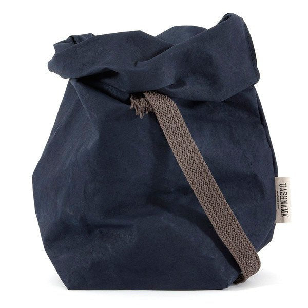 A navy washable paper bag with a single cotton carry strap in brown. The top is displayed open and rolled down, with a natural toned UASHMAMA logo tab on the right hand side.