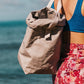A woman is shown walking along a beach carrying a dove grey washable paper bag with two cream cotton straps slung over her shoulder. She wears a blue bikini top and a red printed skirt.