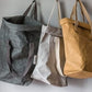 Three washable paper bags are shown hanging from hooks on a grey wall. From left to right they are dark grey, dove grey, and natural tan coloured. All three have the tops rolled down and feature two cotton straps each.