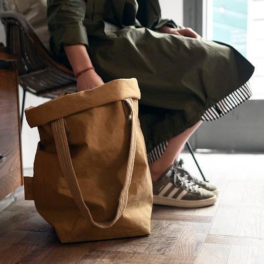 A woman is shown sitting in a chair, wearing a green khaki outfit and green suede sneakers. To her right is a tan coloured washable paper bag with the top rolled down and a natural brown cotton handle. 