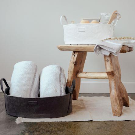 Two washable paper storage baskets, in an oblong shape with side handles, sit atop a rug. The one at left sits on the floor in a black tone, containing two white rolled towels. The one at right sits on a wooden stool, is in a white colour, and features bath products.