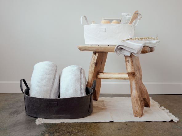 Two washable paper storage baskets, in an oblong shape with side handles, sit atop a rug. The one at left sits on the floor in a black tone, containing two white rolled towels. The one at right sits on a wooden stool, is in a white colour, and features bath products.