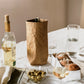 A natural tan washable paper wine holder sits atop a linen table cloth, containing a bottle of red wine. Another bottle of wine sits at left, alongside olives and a water carafe. A washable paper breadbasket containing three bread rolls sits at right.