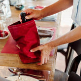 A person is shown removing a bottle of wine from a red washable paper wine holder, which sits atop a laid table.